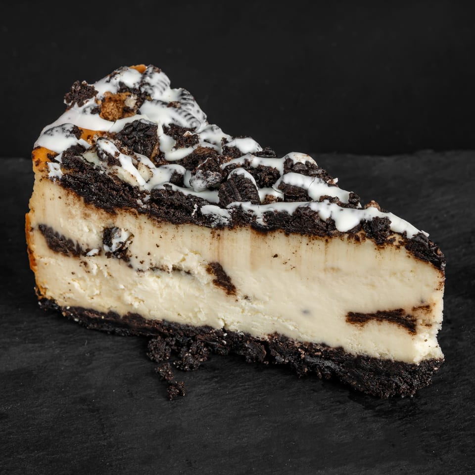 A slice of Drizzle Cafe's cookies and cream cheesecake with white icing drizzle on a dark surface.